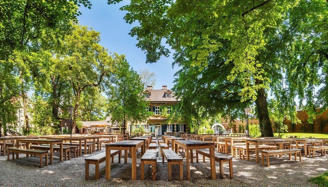 Tavoli e panche in legno vuoti disposti su un patio in ghiaia, sotto grandi alberi verdi e frondosi, di fronte a un edificio.