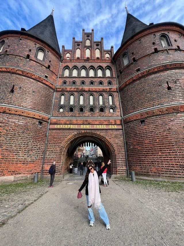 Una donna posa con il segno della pace davanti a un imponente portale medievale in mattoni, con due torri a tetto conico, sotto un cielo azzurro.