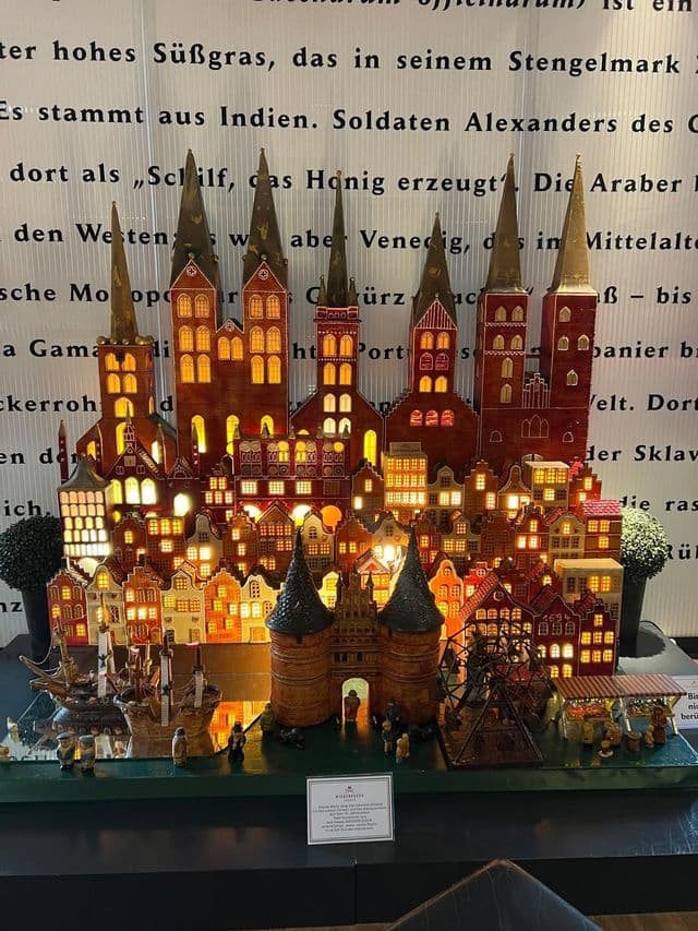 Un modellino in miniatura illuminato di una città storica con chiese, case, una porta cittadina e navi, esposto su uno sfondo testuale.