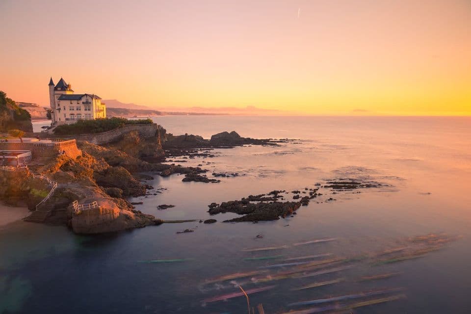 Eine Villa auf einer felsigen Klippe überblickt bei Sonnenuntergang das Meer, mit bunten Langzeitbelichtungsspuren von Booten im Wasser.