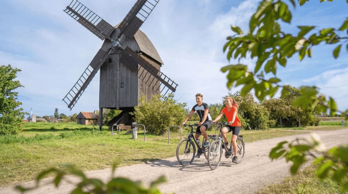 Ein Paar fährt auf einem Schotterweg auf dem Land Fahrrad, mit einer großen hölzernen Windmühle im Hintergrund.