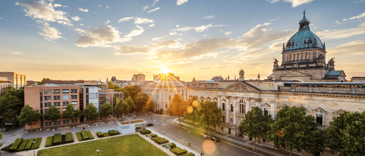 Eine Luftaufnahme eines verzierten historischen Gebäudes mit einer grünen Kuppel in einer Stadtlandschaft bei Sonnenuntergang, mit einem Park im Vordergrund.