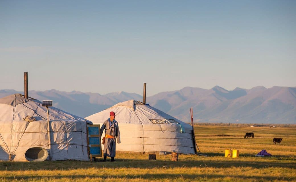 Una persona con atuendo tradicional se encuentra entre dos yurtas en una llanura herbosa con montañas distantes.