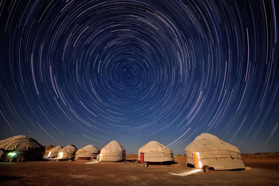 Una fila de yurtas iluminadas en un campamento en el desierto bajo un cielo nocturno con rastros de estrellas circulares de una exposición prolongada.