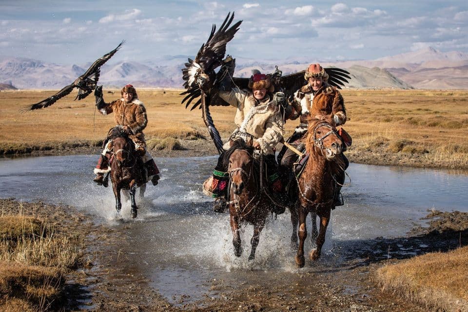 Tres cazadores de águilas con vestimenta tradicional de piel montan a caballo a través del agua en una vasta llanura, sosteniendo águilas con las alas extendidas.