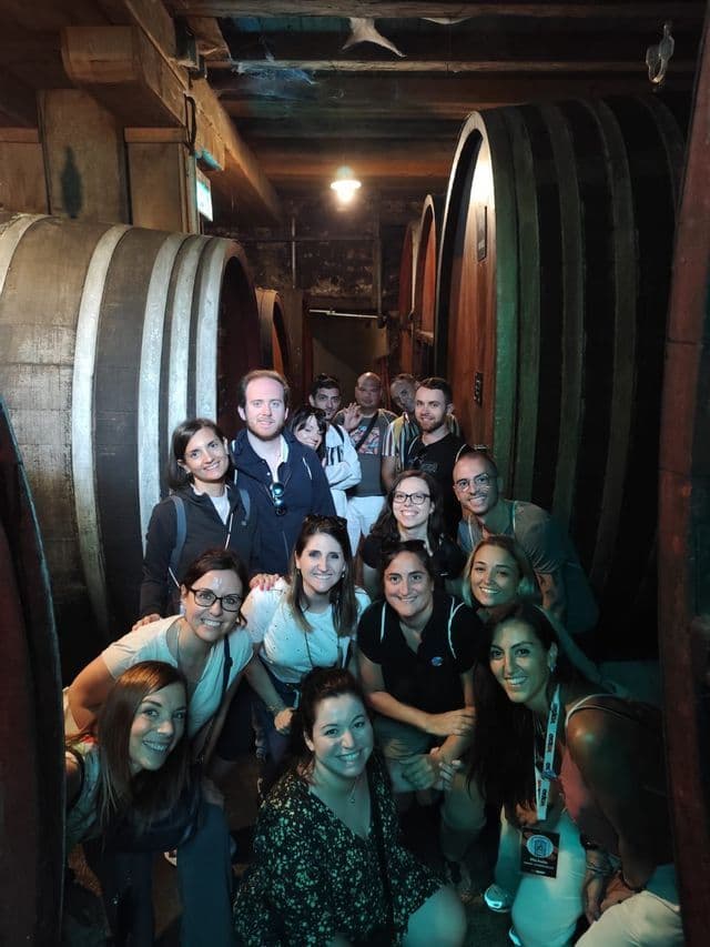 Un gruppo WeRoad si mette in posa per una foto all'interno di una cantina, in piedi tra grandi botti di legno.