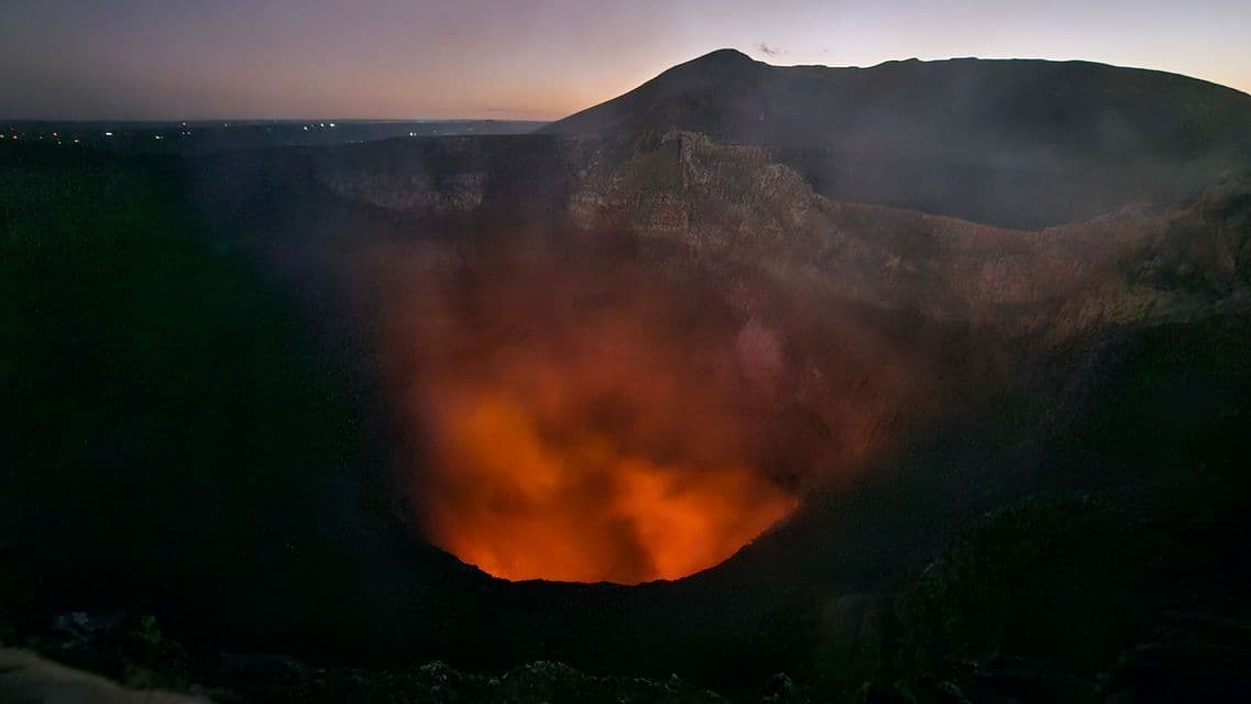 Ein Vulkankrater glüht in der Dämmerung mit orangefarbener, geschmolzener Lava, und Rauch steigt in den Zwielichthimmel auf.