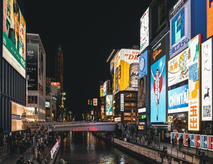 Un bullicioso canal urbano por la noche, con gente paseando por las orillas bajo enormes carteles de neón luminosos.