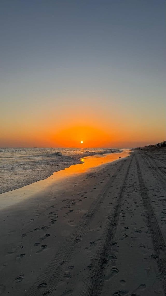 Il sole tramonta sull'oceano, proiettando un riflesso dorato su una spiaggia sabbiosa coperta di impronte e tracce di pneumatici.