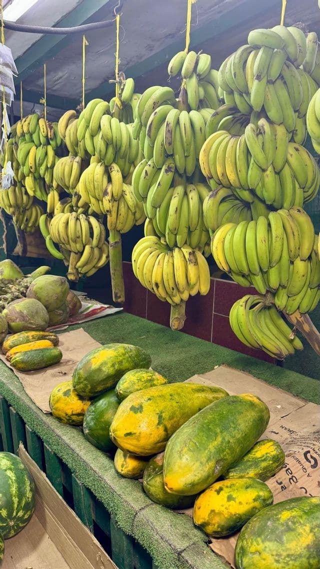 Caschi di banane verdi e gialle appesi sopra un espositore di papaya fresche e altri frutti in una bancarella del mercato coperto.