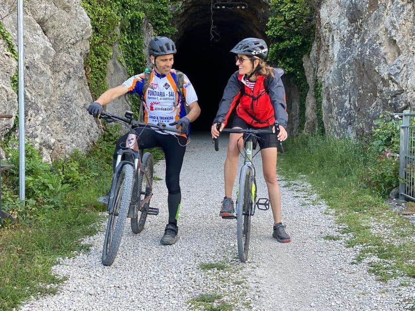 Due ciclisti con caschi si sorridono, in sosta sulle bici su un sentiero sterrato che conduce a un tunnel buio.