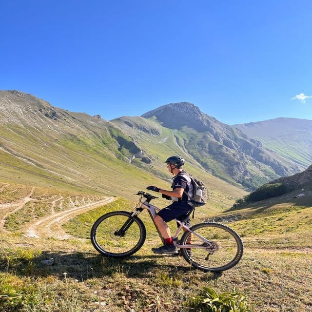 Un ciclista con casco in mountain bike si ferma su un sentiero erboso con vista su una catena montuosa sotto un cielo sereno.