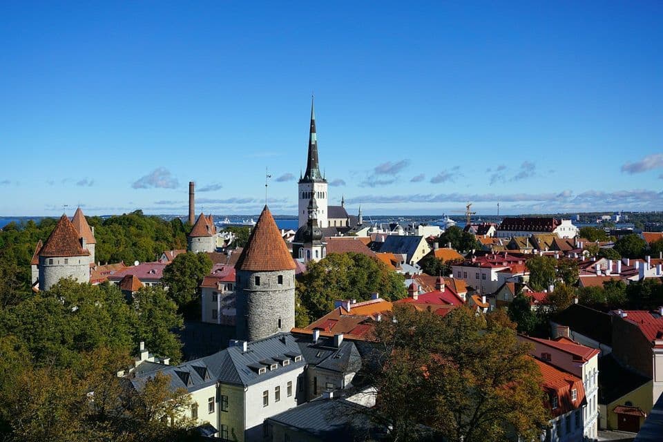 Un paesaggio urbano di una città storica con torri in pietra, una guglia di chiesa e tetti rossi sotto un cielo azzurro brillante.