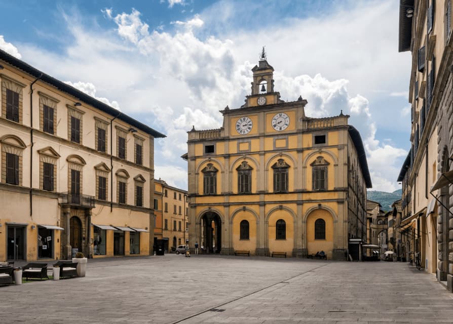 Un grande edificio giallo con una torre dell'orologio si erge alla fine di una spaziosa piazza pavimentata sotto un cielo azzurro nuvoloso.