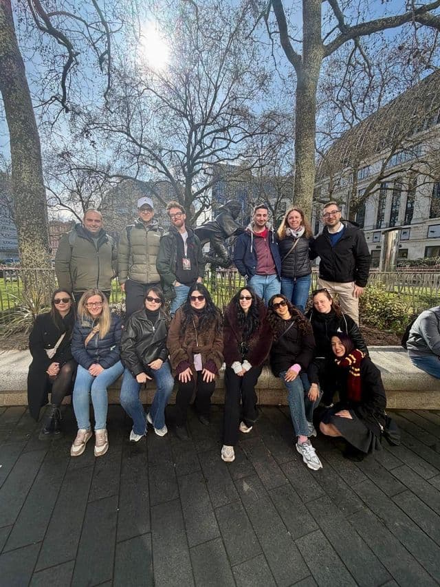 Un gruppo WeRoad in viaggio posa per una foto in un parco urbano soleggiato, con alcuni partecipanti seduti su un muretto di pietra e altri in piedi alle loro spalle.