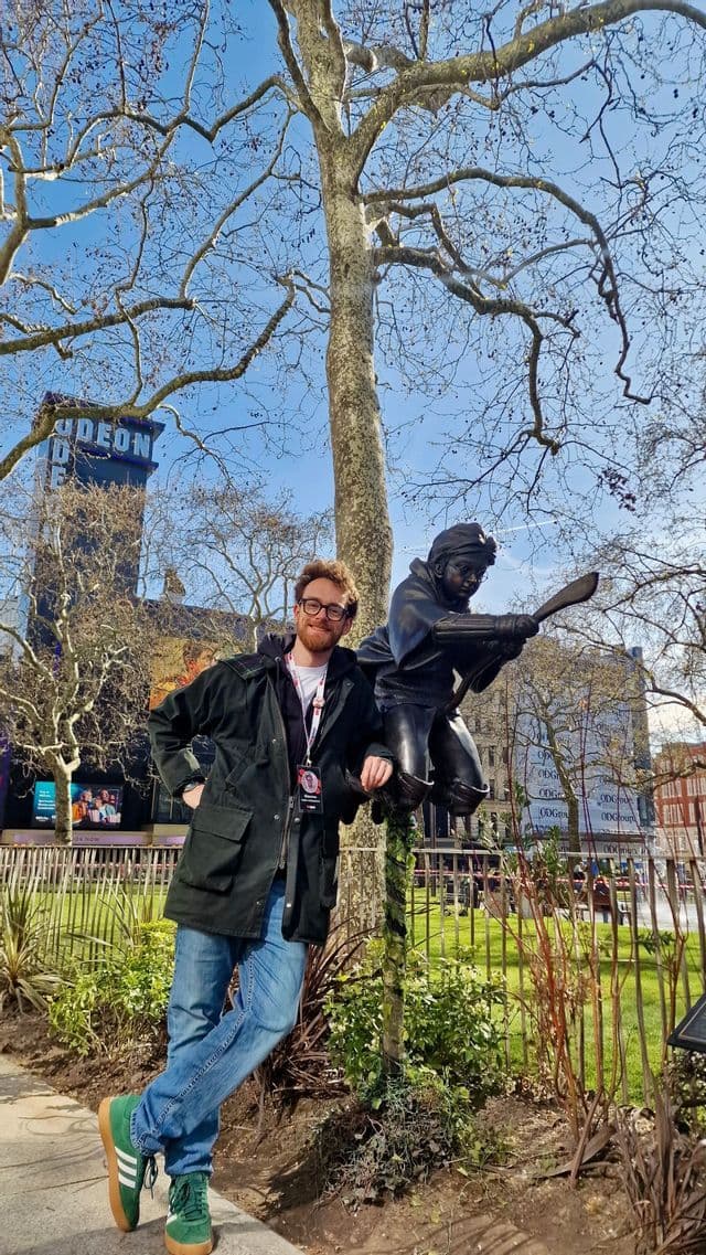 Un uomo con gli occhiali posa accanto a una statua di bronzo di Harry Potter su una scopa in un parco cittadino in una giornata di sole.