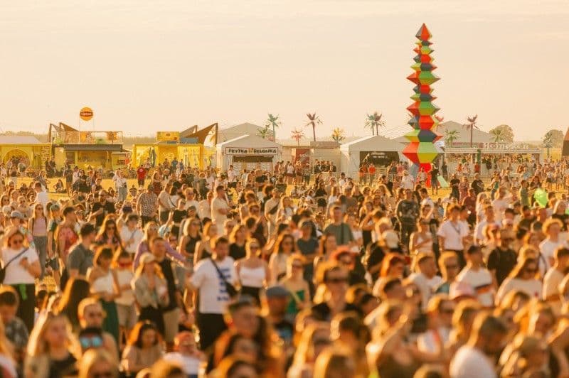 Una grande folla di persone si è radunata a un festival all'aperto, con tende e una scultura alta e colorata sullo sfondo sotto un cielo fosco.