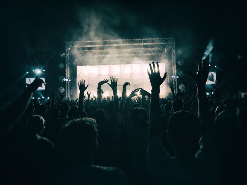 Una folla di persone in silhouette con le mani alzate davanti a un palco illuminato a giorno durante un concerto notturno.
