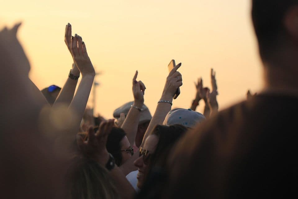 Una folla di persone con le mani alzate a un evento all'aperto, sullo sfondo di un cielo al tramonto.