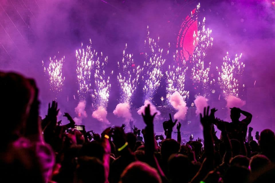 Una folla in silhouette alza le mani verso un palco con fuochi d'artificio e fumo viola durante un festival musicale.