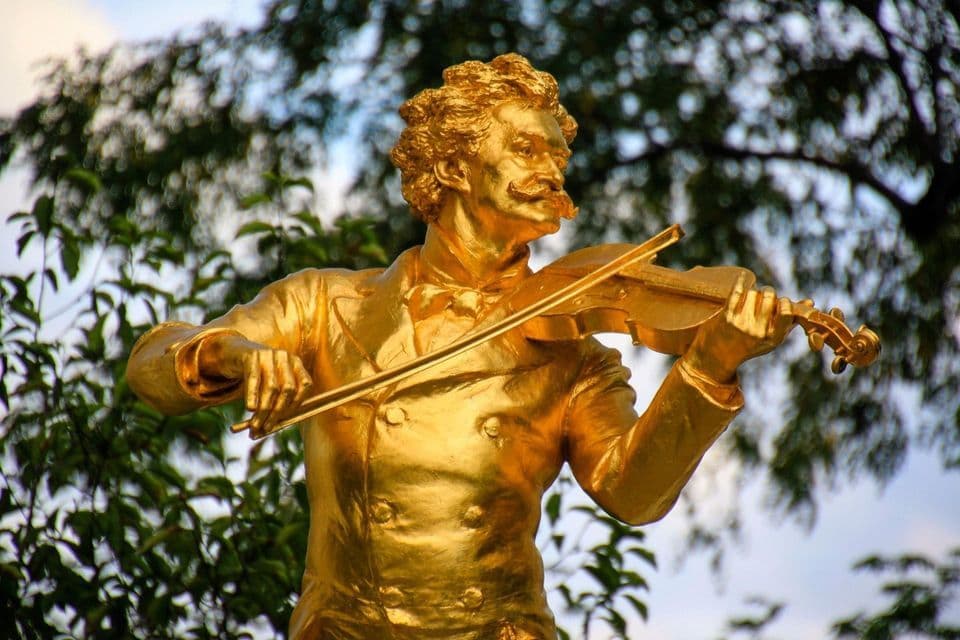 Une statue dorée d'un homme moustachu jouant du violon, avec des arbres verts flous en arrière-plan.
