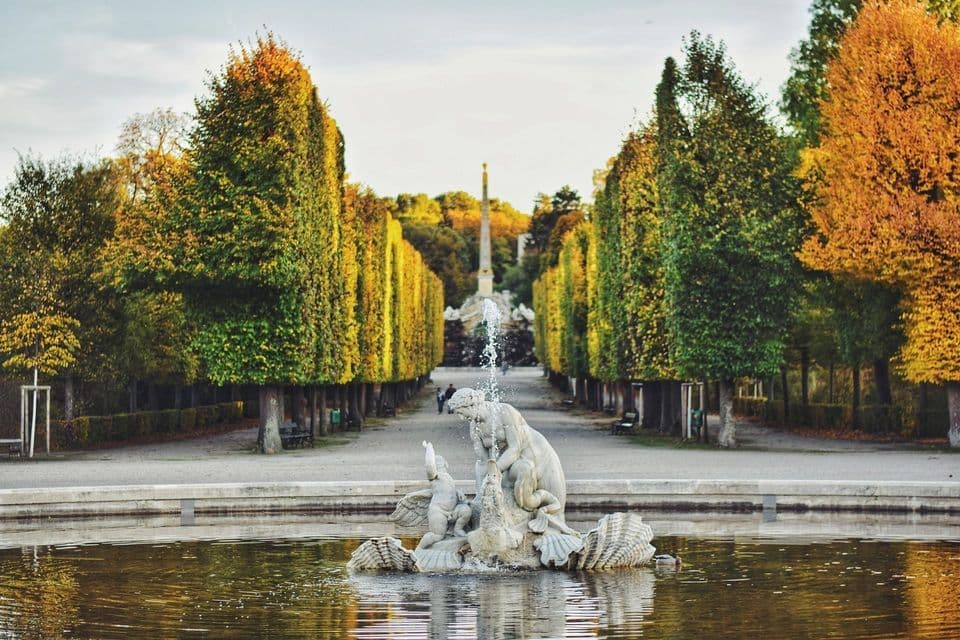 Une fontaine centrale avec une statue en marbre est située dans un bassin, faisant face à une longue allée d'arbres taillés aux couleurs automnales.