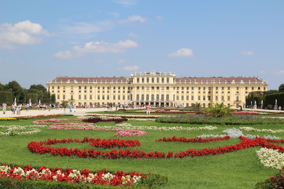 Un grand palais jaune se dresse derrière des jardins bien entretenus avec des parterres de fleurs colorés sous un ciel bleu parsemé de nuages.