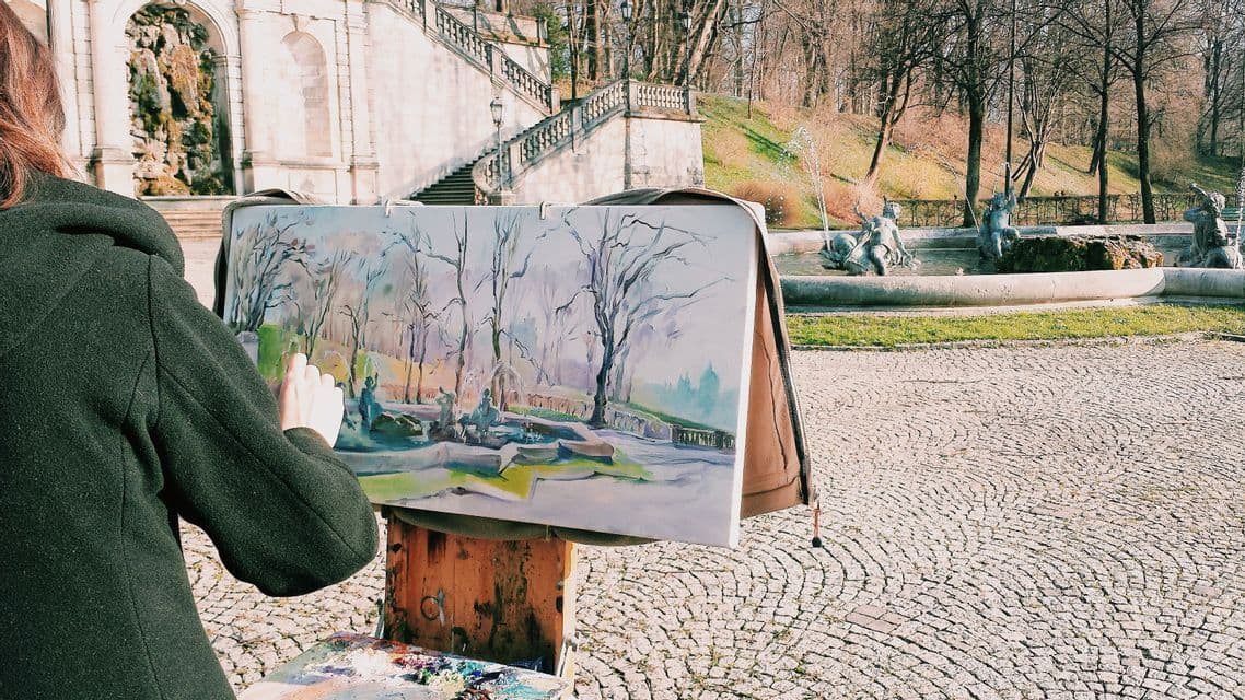 Vue par-dessus l'épaule d'un artiste peignant une scène de parc avec une fontaine et des arbres sur une toile en plein air.