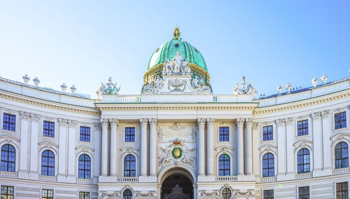 La façade blanche et ornée d'un grand palais, avec un grand dôme vert, des colonnes et des statues, se détachant sur un ciel bleu vif.