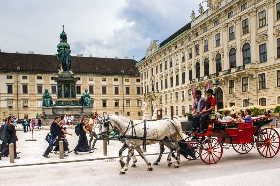 Deux chevaux gris pommelés tirent une calèche touristique rouge sur une place de ville animée, devant un bâtiment historique orné et un monument.