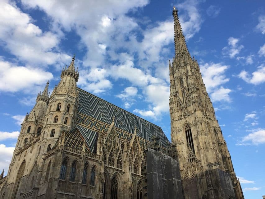 Une cathédrale gothique ornée, avec un toit en tuiles colorées et à motifs et une haute flèche, vue en contre-plongée sur un ciel nuageux.