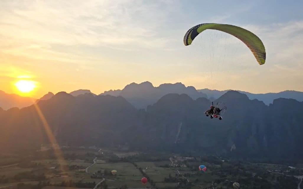 Un parapendio a motore sorvola una valle di montagna all'alba, con diverse mongolfiere visibili sotto.