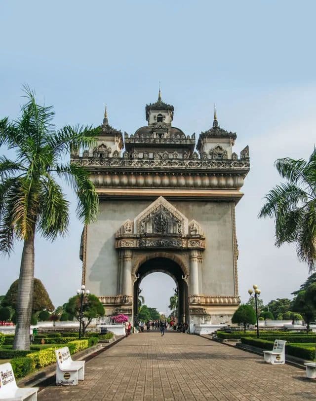 Un monumento ad arco in pietra riccamente decorato si erge in un parco, con palme e un viale pavimentato che vi conduce sotto un cielo sereno.