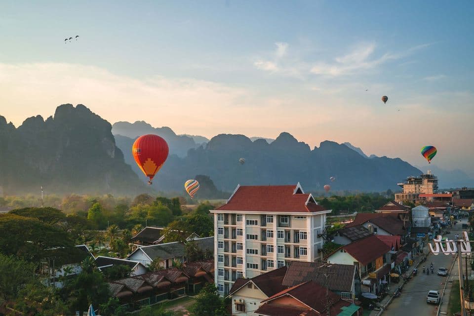 Diverse mongolfiere fluttuano sopra una città ai piedi di grandi montagne verdi all'alba, con parapendio nel cielo.