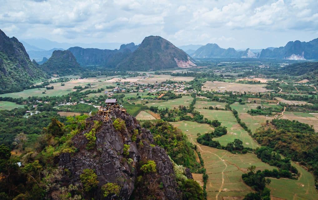 Una vista aerea di un viaggio di gruppo WeRoad su un belvedere in legno, in cima a un picco roccioso, che domina una vasta valle verde e le montagne.
