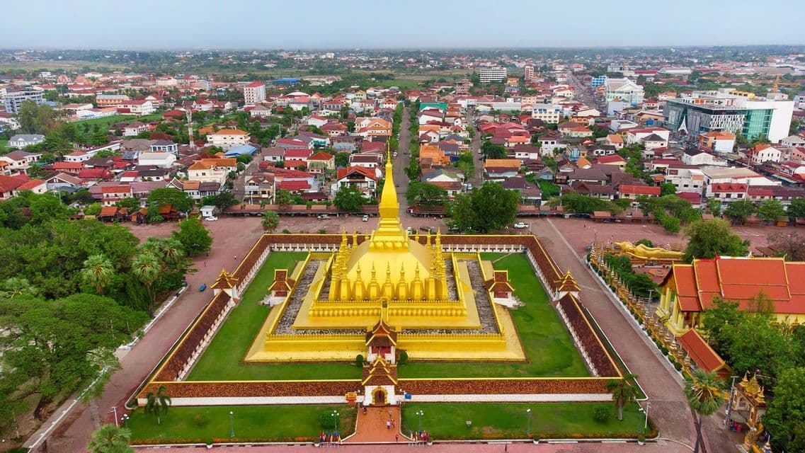 Veduta aerea di una grande stupa dorata in un cortile recintato, con una vasta città sullo sfondo sotto un cielo coperto.