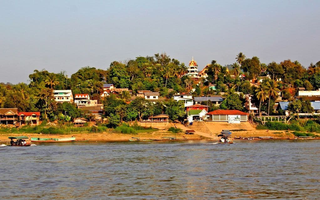 Piccole barche scorrono su un fiume, oltre un villaggio collinare con alberi lussureggianti e un tempio.