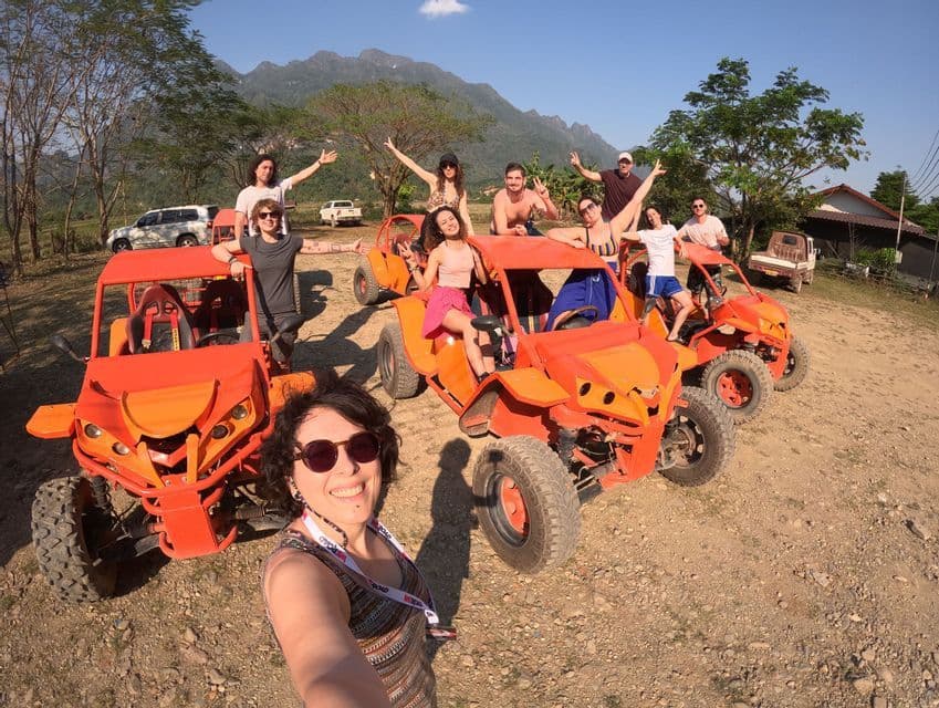 Un gruppo WeRoad si scatta un selfie posando su e attorno a vivaci buggy arancioni, con uno sfondo di paesaggio rurale e montagne.