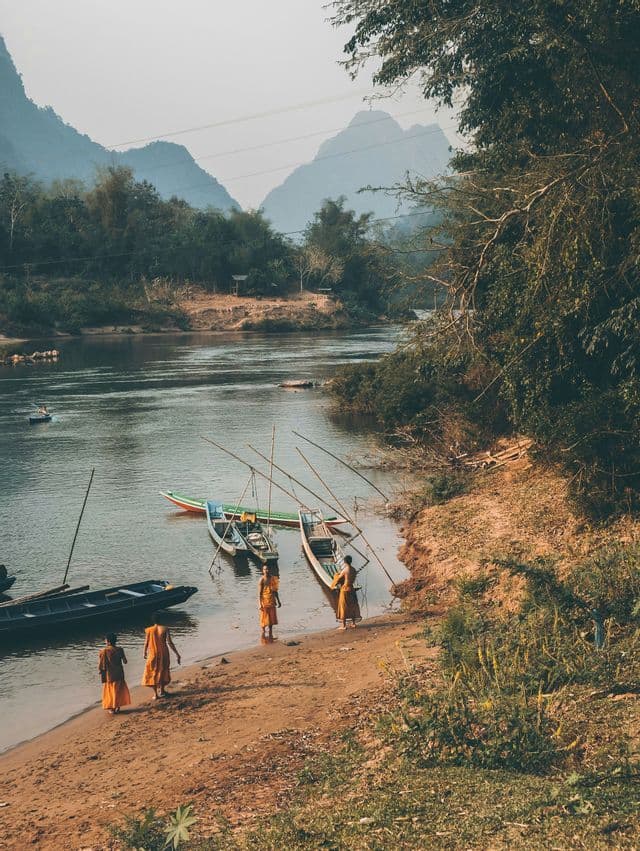 Quattro monaci con tuniche arancioni camminano lungo la riva sabbiosa di un fiume verso lunghe barche ormeggiate in acqua.