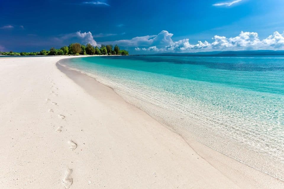Una spiaggia di sabbia bianca con impronte si curva lungo l'oceano turchese trasparente sotto un cielo blu con nuvole soffici.