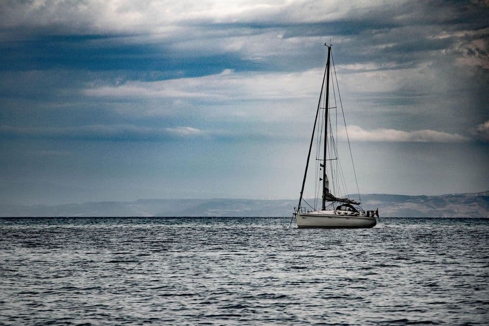 Una barca a vela bianca galleggia su acque agitate sotto un cielo nuvoloso e drammatico, con colline lontane all'orizzonte.