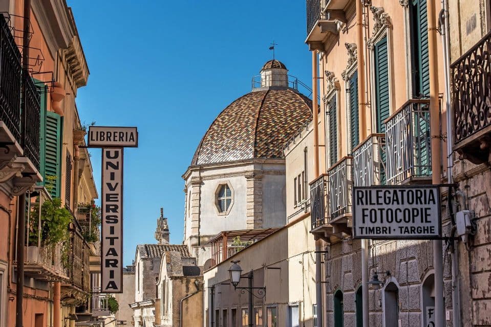 Una strada stretta fiancheggiata da vecchi edifici e insegne italiane, con una cupola piastrellata visibile contro un cielo azzurro chiaro.