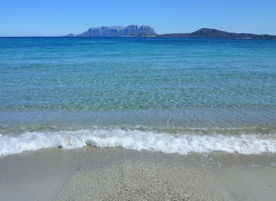 Acque cristalline turchesi lambiscono una spiaggia sabbiosa, con una grande isola rocciosa visibile all'orizzonte sotto un cielo azzurro.