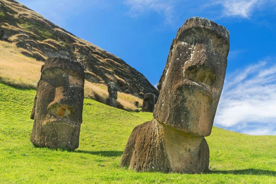 Diverse grandi statue Moai di pietra si ergono su una collina erbosa sotto un cielo azzurro brillante.