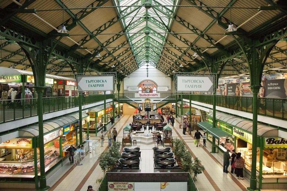 Das Innere einer zweistöckigen Markthalle mit grünen Metallbögen und einem Glasdach, in der Menschen durch Geschäfte bummeln und in Cafés sitzen.