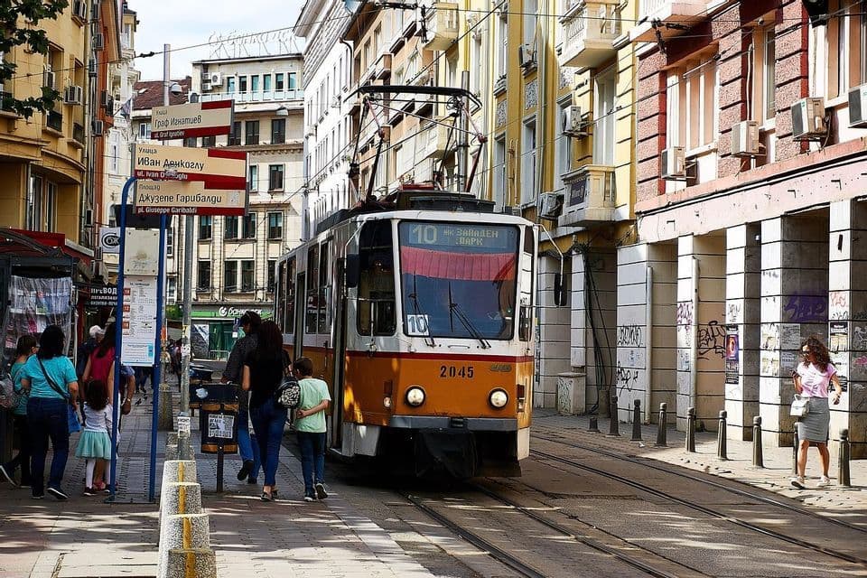 Eine gelb-weiße Straßenbahn fährt auf Schienen eine enge Stadtstraße entlang, mit Menschen, die auf dem Bürgersteig daneben gehen.