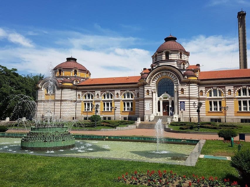 Ein großes, verziertes Gebäude mit gestreiften Wänden und Kuppeln hinter einem öffentlichen Park mit einem gestuften grünen Springbrunnen, der Wasser sprüht.