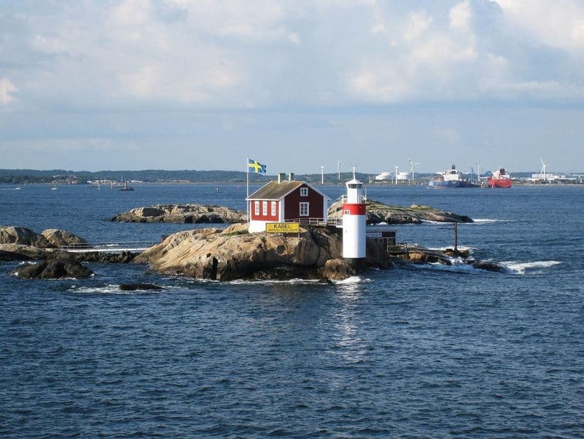 Ein rotes Holzhaus und ein kleiner Leuchtturm stehen auf einer Felseninsel im Meer, mit wehender schwedischer Flagge.