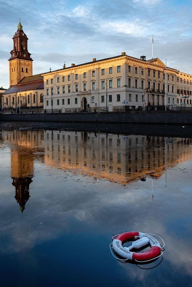 Ein klassisches Gebäude und ein Kirchturm spiegeln sich im ruhigen Kanalwasser wider, im Vordergrund schwimmt ein Rettungsring.