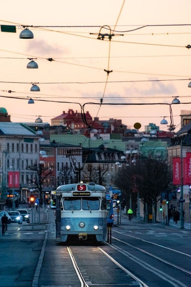 Eine hellblau-weiße Straßenbahn fährt in der Dämmerung mit eingeschalteten Scheinwerfern auf einer Stadtstraße unter einem Netz von Oberleitungen entlang.